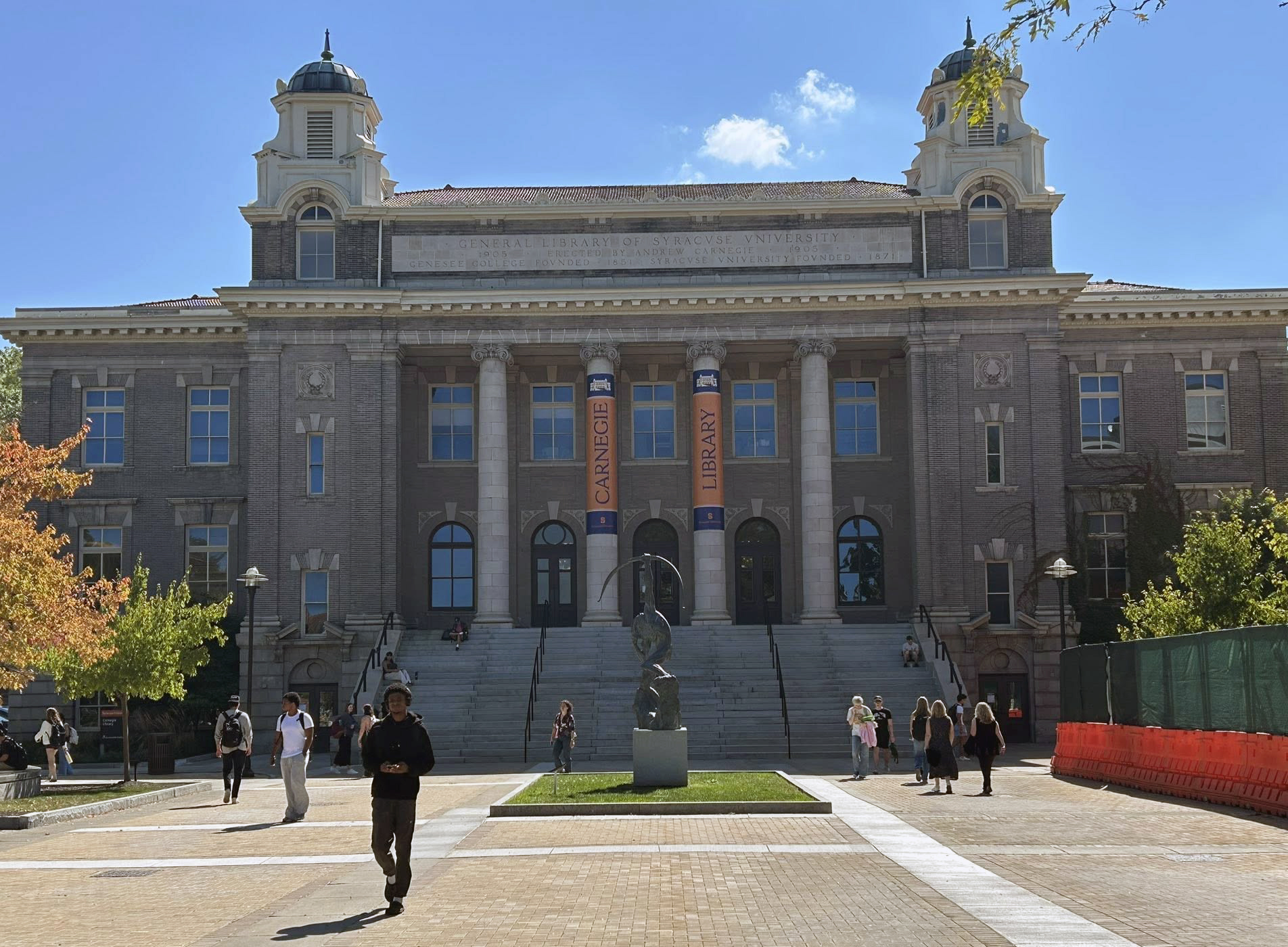 Front view of Carnegie Library at Syracuse University, with twin towers, columned entrance, courtyard sculpture and people walking nearby.
