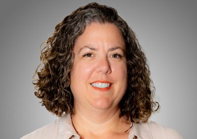 A professional headshot of a woman with shoulder-length curly brown hair with gray highlights, wearing a light-colored blouse and smiling warmly at the camera against a neutral gray background.