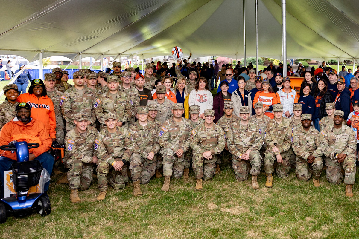 A large group of military personnel in camouflage uniforms and Syracuse University community members gather for a group photo under a white tent at an outdoor event, with many wearing Syracuse orange clothing and merchandise visible throughout the crowd.