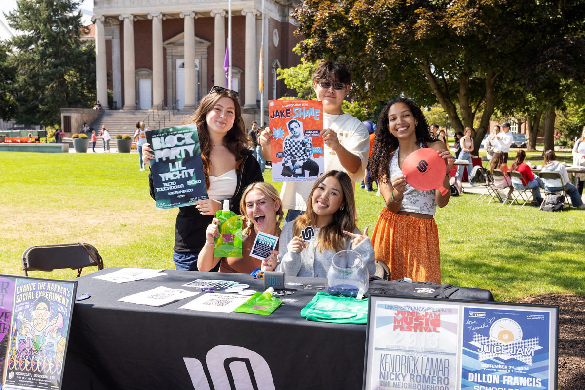 Five students posing with promotional materials and posters at a campus organization table, with a classical columned building in the background during an outdoor student activities fair.