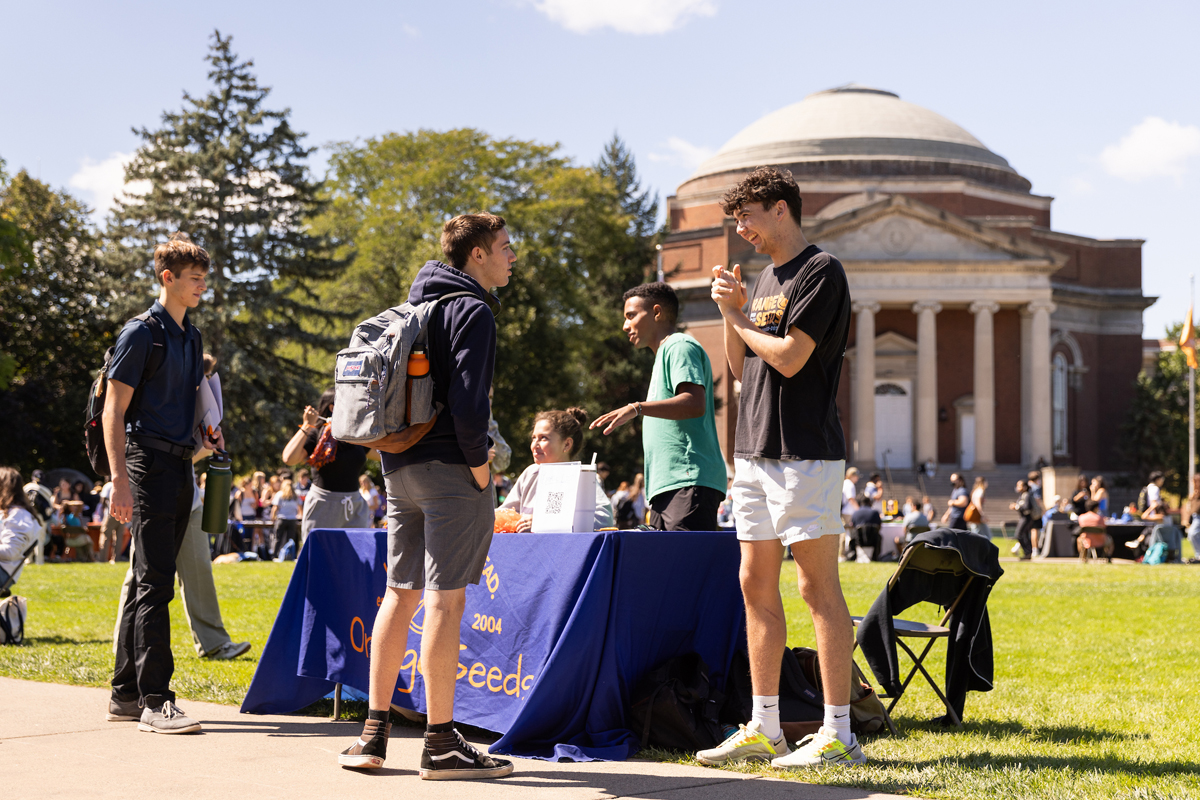 Students gathering around a blue information table on a college campus quad, with a domed academic building and trees in the background on a sunny day.