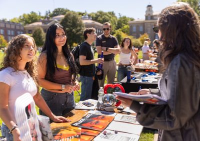 Students browsing informational materials at an outdoor campus fair or club event, with tables displaying brochures and promotional items on a sunny day.
