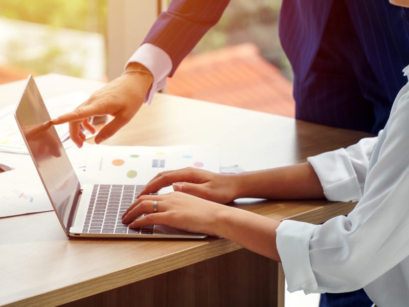 Two professionals collaborating at a laptop on a wooden desk with charts and documents visible in bright natural lighting.