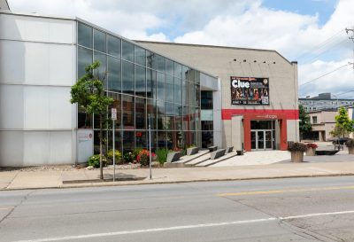 Modern building with large glass windows and a "Clue" event sign dated June 7-25 above the entrance, surrounded by greenery.