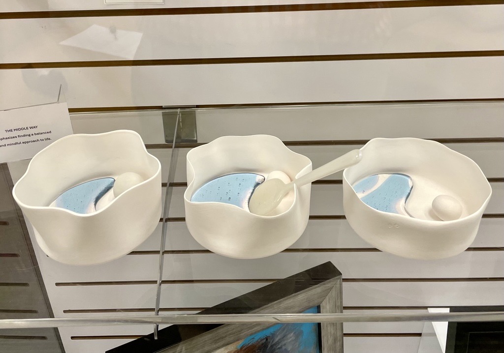 Three white ceramic bowls with wavy edges and blue abstract designs, displayed on a glass shelf; one bowl contains a white spoon.