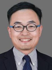 A person wearing a dark suit, checkered shirt, and blue tie with white dots and glasses. 