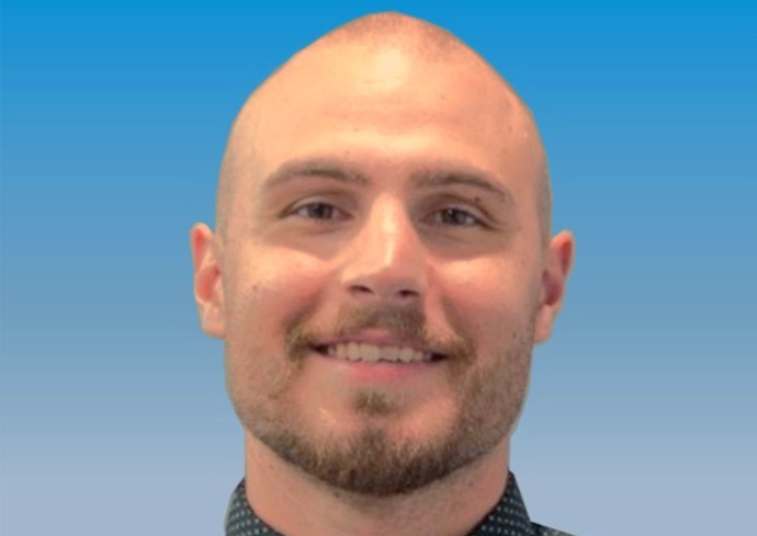 A professional headshot of a smiling man with a goatee wearing a dark patterned shirt against a blue gradient background.