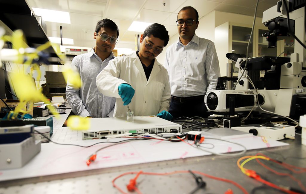 A person in a lab coat and gloves works with equipment at a table covered covered with wires, electronics and a microscope, while two people observe.