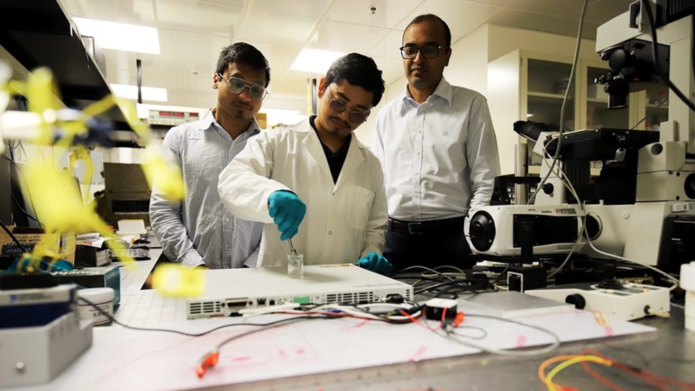 One person in a lab coat and gloves works with equipment at a table covered with wires, electronics and a microscope, while two other people observe.