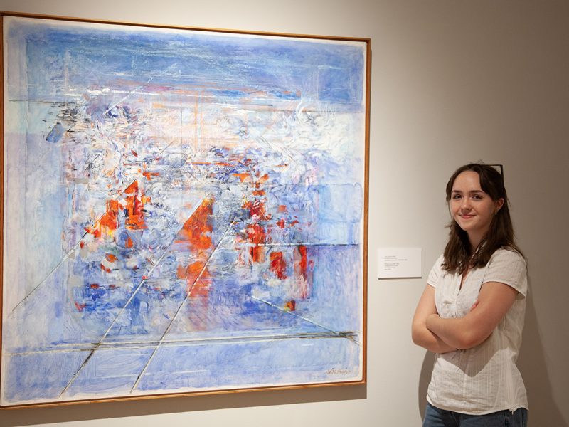 A smiling person with long dark hair in a white top stands with crossed arms next to a large abstract painting featuring blue, white, and orange brushstrokes in a gallery setting.