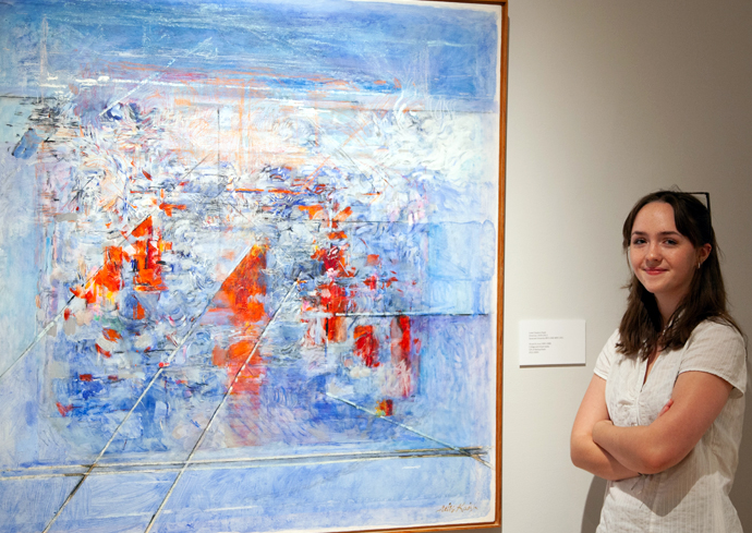 A smiling person with long dark hair in a white top stands with crossed arms next to a large abstract painting featuring blue, white, and orange brushstrokes in a gallery setting.