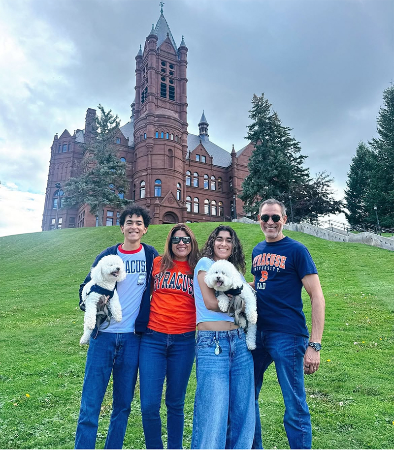 four people in Syracuse University apparel holding two small white dogs, stand on a grassy hill in front of a historic red-brick campus building.