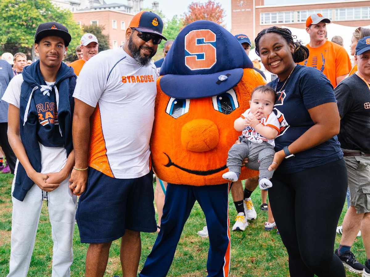 Group photo with Syracuse University mascot Otto the Orange on campus lawn, surrounded by people in Syracuse apparel, including a baby in themed clothing.