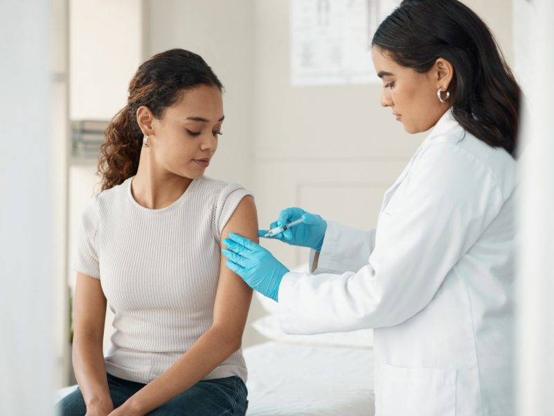 Healthcare professional administering a vaccine to a seated person in a clinical exam room.