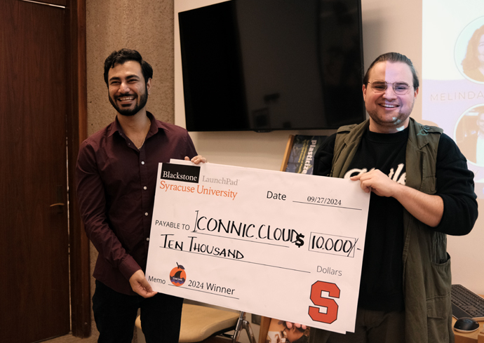 Two students smiling and holding a large ceremonial check made out to "Iconic Clouds" for $10,000 as the "2024 Winner." The man on the left wears a burgundy button-up shirt, while the man on the right wears a black t-shirt and olive jacket.