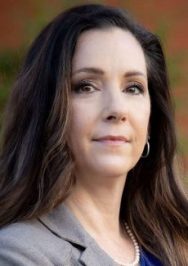 A person with long, wavy hair wearing a gray blazer, blue shirt, and pearl necklace stands outdoors.