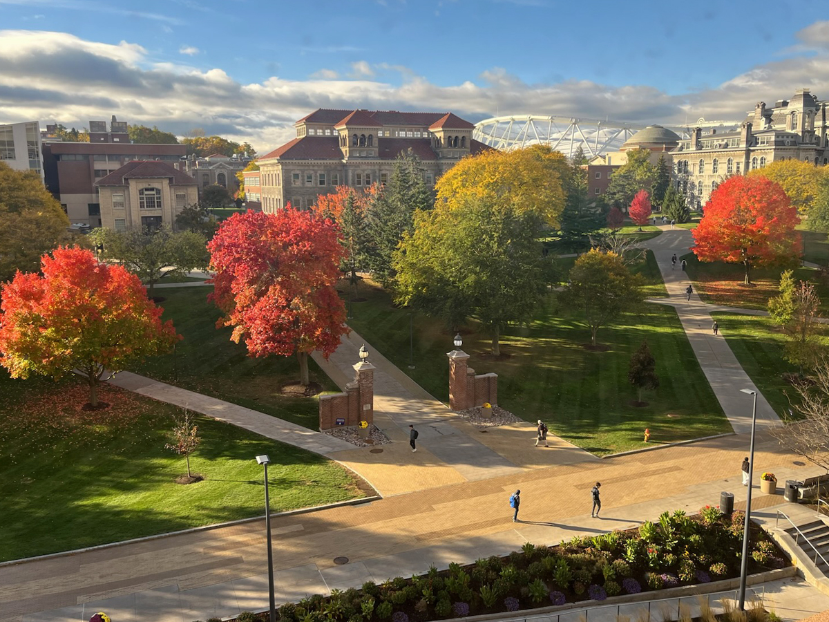 Aerial view of campus quadrangle with vibrant red and yellow fall foliage, brick gateway pillars, walking paths and historic buildings under blue sky.