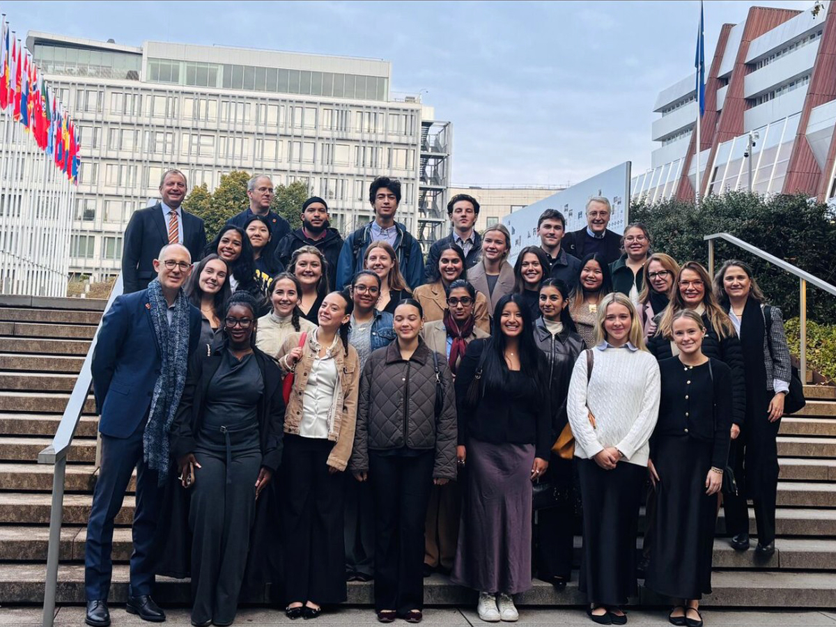 A large group of Syracuse Abroad students and faculty posing on outdoor steps in front of modern office buildings with international flags visible in the background at the Council of Europe.