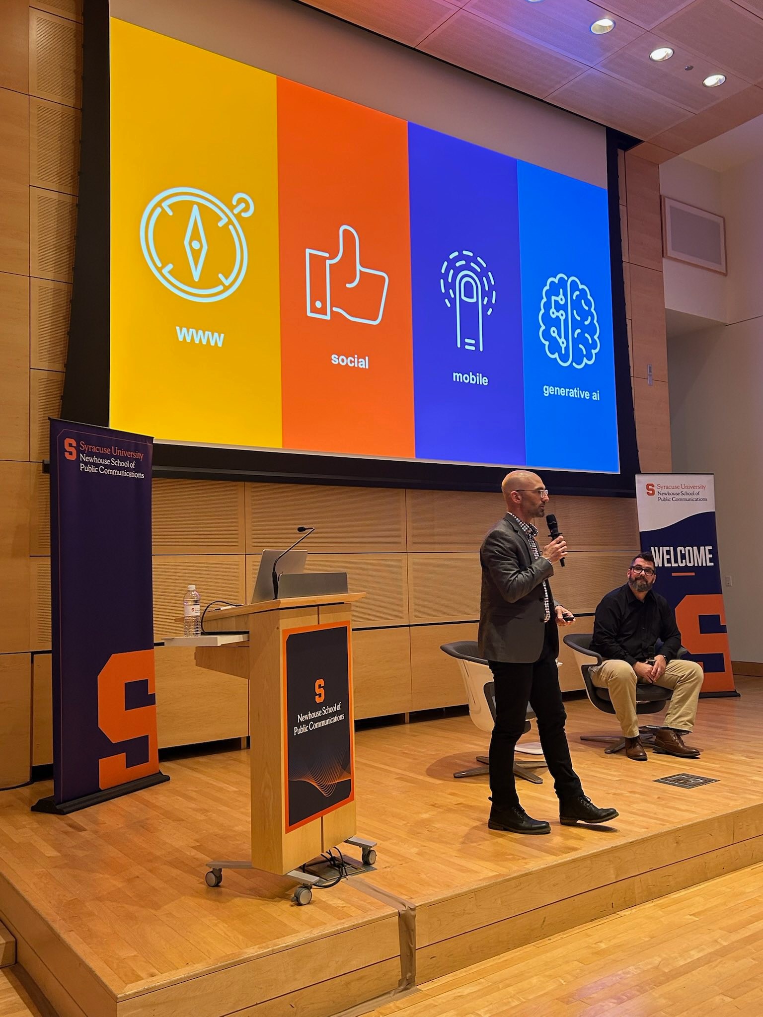 Speaker presenting at Newhouse School podium with colorful slide showing evolution of digital media: www, social, mobile, and generative AI icons