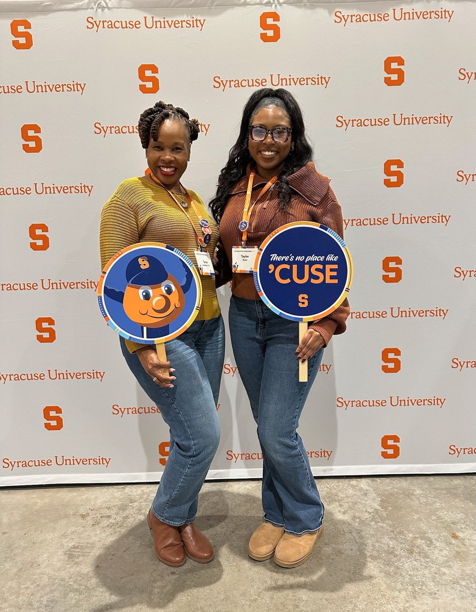 Two people posing at Syracuse University branded backdrop, holding circular signs featuring Otto mascot and "There's no place like 'CUSE" message