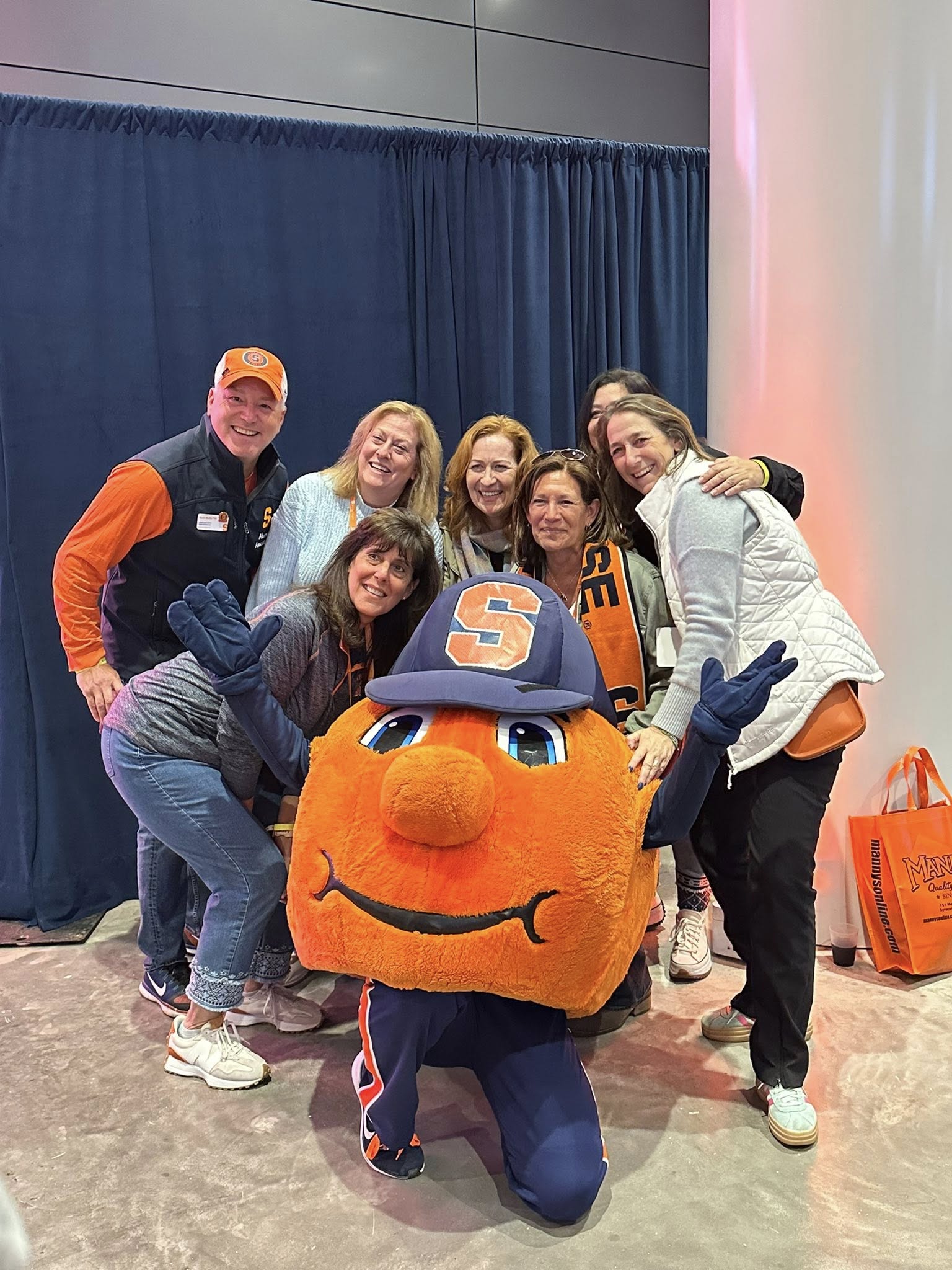 Group of alumni gathered around Otto the Orange mascot in front of blue curtain backdrop during Homecoming event