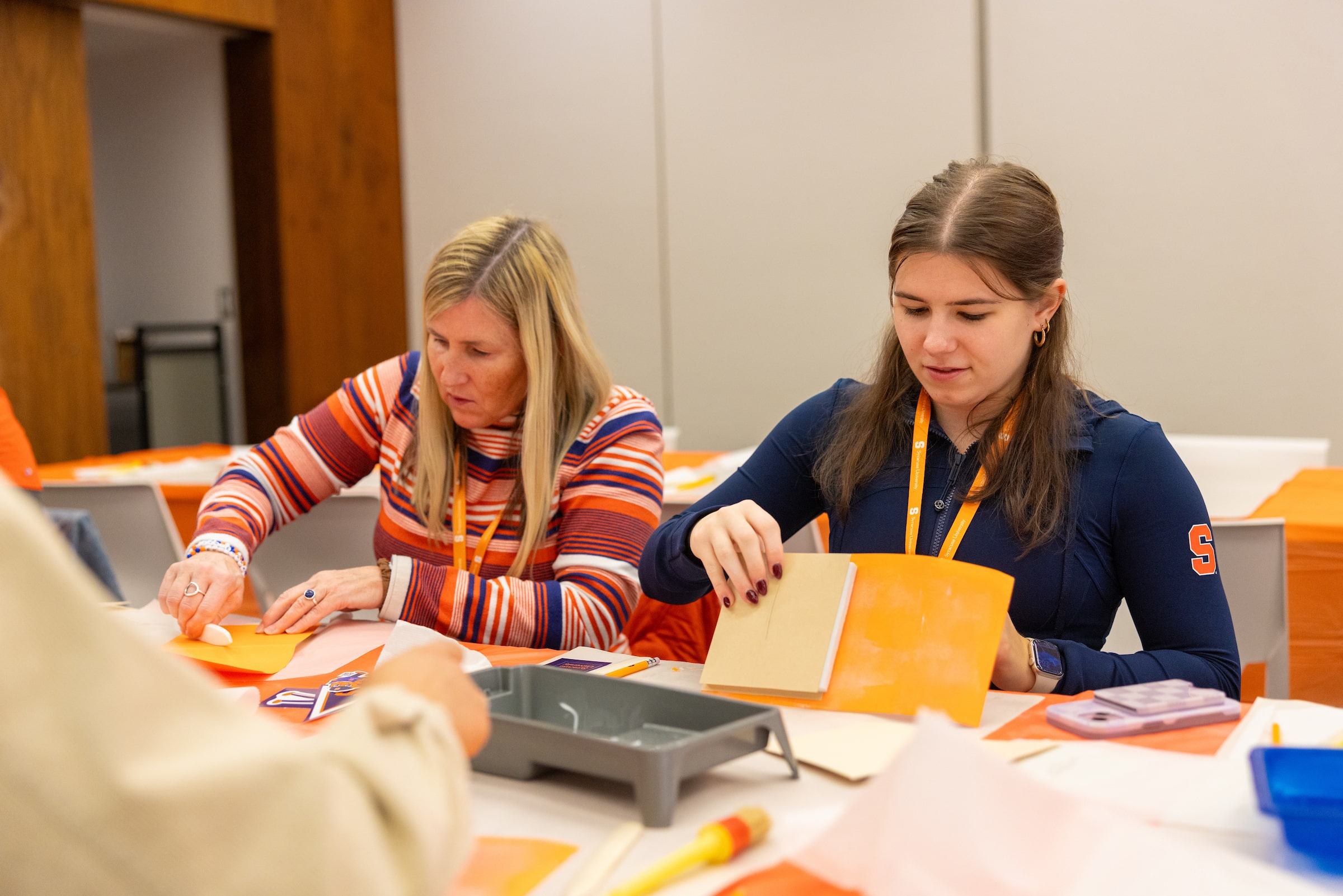 Two people collaborate at a table, working with orange folders and office supplies in a bright meeting room