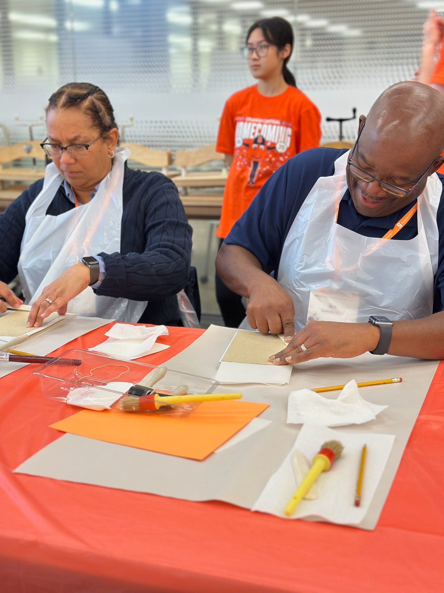 Alumni participants wearing white aprons engaged in arts and crafts activity at table with orange and white materials