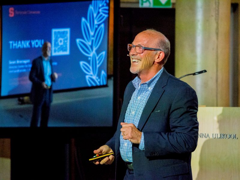 Speaker presenting at a conference with a large screen behind displaying a thank-you slide featuring text and a decorative leaf graphic