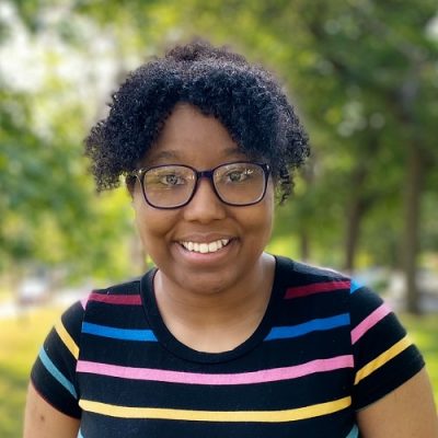 Person wearing a bright red sweater with straight, shoulder-length dark hair, standing outdoors in front of a light-colored stone wall and greenery.