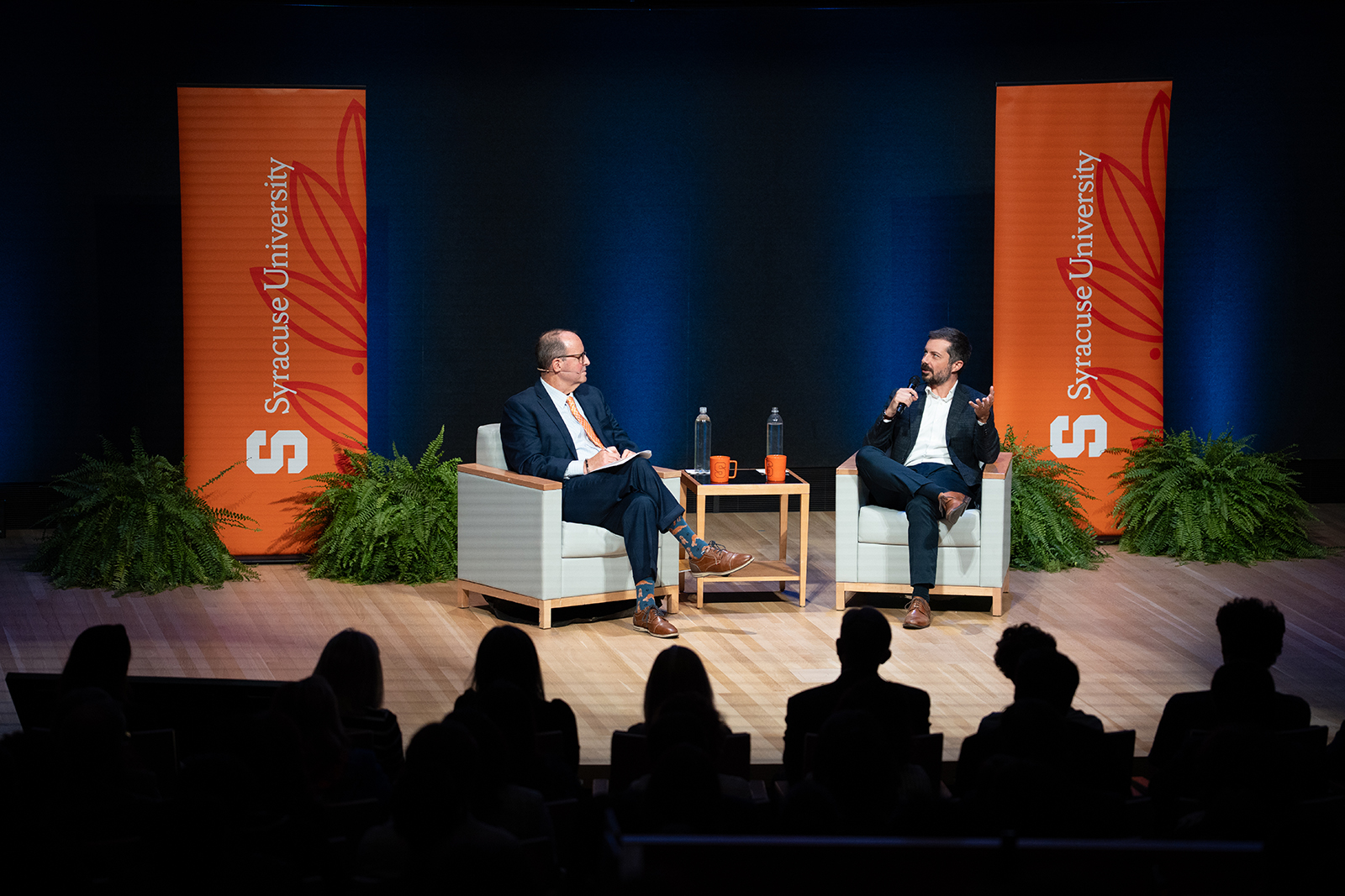 Two people sitting on stage in a discussion at a Syracuse University event, with audience in the foreground and a backdrop featuring the university's logo