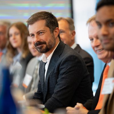 Group of professionals, including former US Transportation Secretary Pete Buttigieg, sitting at a conference table, engaged in a discussion.