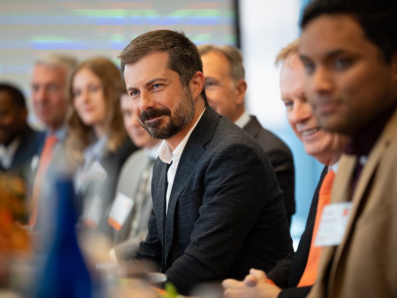 Group of professionals, including former US Transportation Secretary Pete Buttigieg, sitting at a conference table, engaged in a discussion.