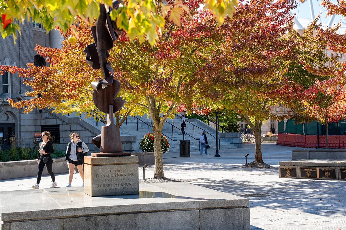 Abstract metal sculpture on Syracuse University campus plaza, surrounded by autumn trees and pedestrians