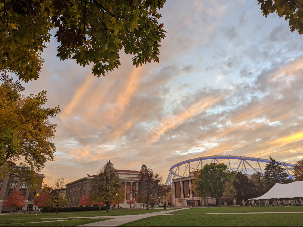 The quad is pictured alongside the JMA Wireless Dome and Carnegie Library under a dramatic sunset sky with orange and pink clouds framed by fall foliage.