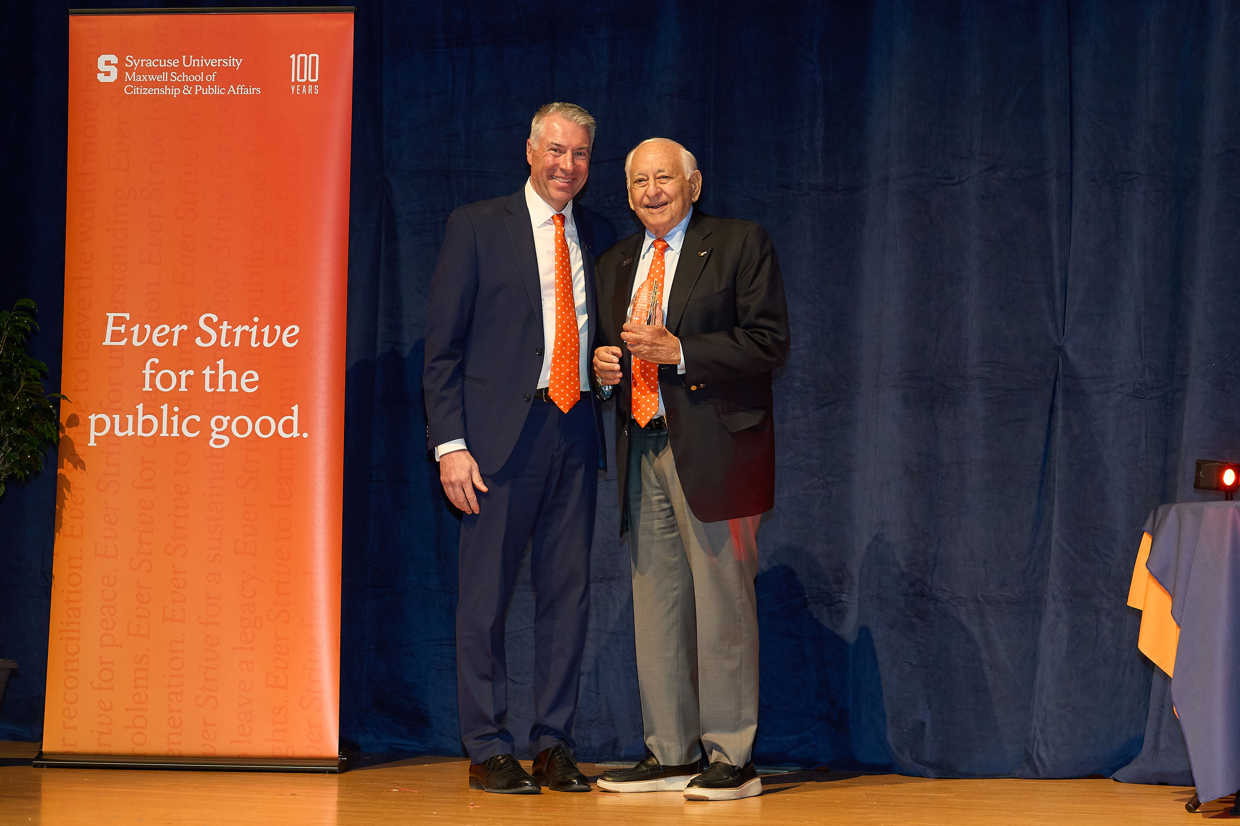 Two people in formal attire stand on a stage with a blue curtain and an orange Syracuse University Maxwell School banner.
