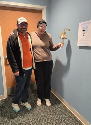 Two people stand in a hallway as one rings a bell beneath a 'Ringing for Hope Against Cancer' sign.