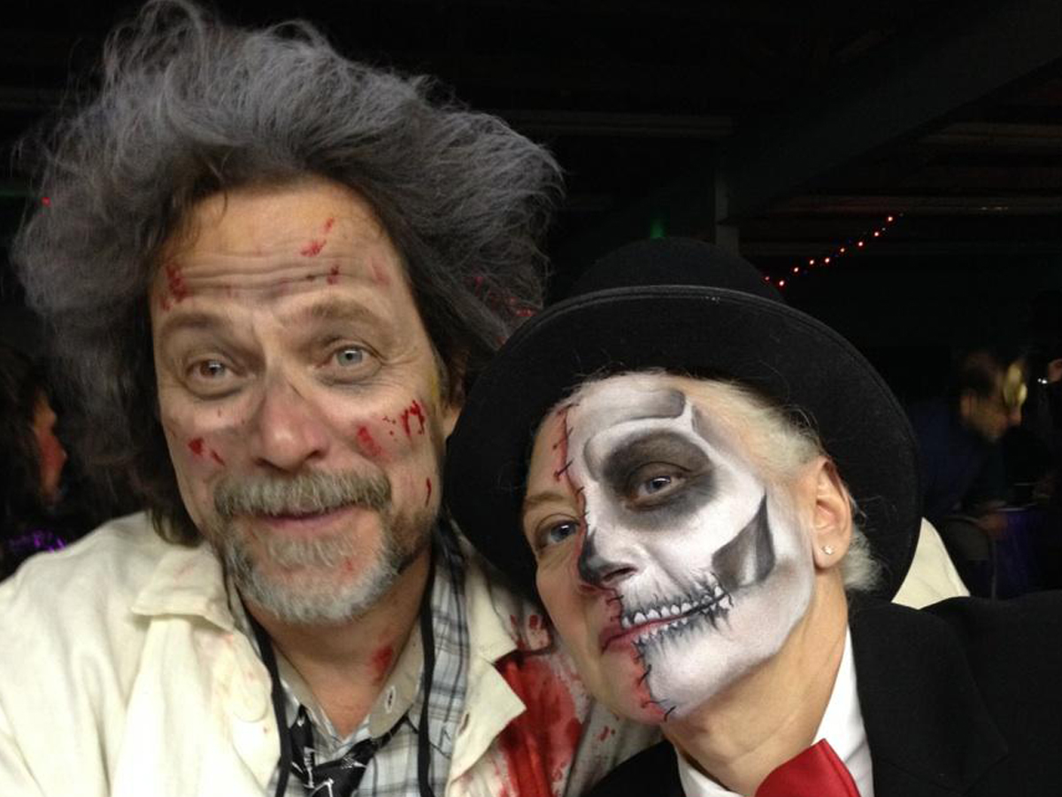 Two people in elaborate Halloween costumes with theatrical makeup - one wearing wild, voluminous hair with fake blood and wounds, the other in a black top hat with skull face paint featuring skeletal teeth and shading.