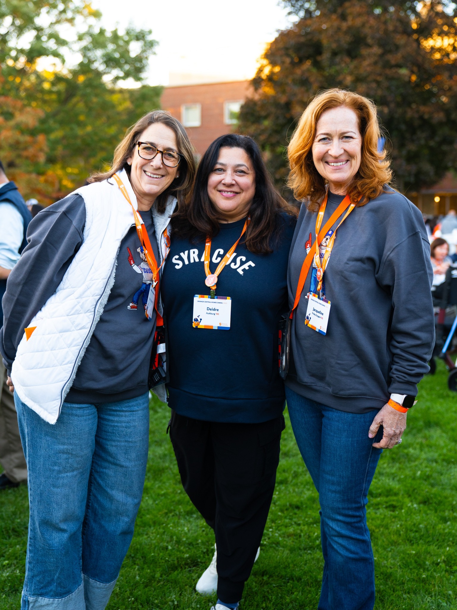 Three alumni wearing Syracuse apparel and orange lanyards pose together outdoors on campus at dusk