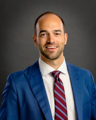 Person wearing a blue suit jacket, white dress shirt, and red striped tie against a dark gray background.