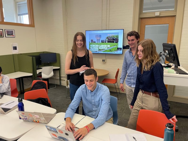 Four people collaborate around a laptop in a modern classroom; a screen behind them displays “Forced Labor in Global Supply Chains.”