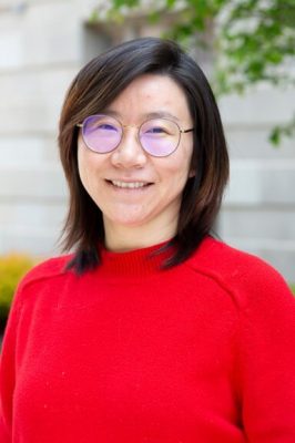 Person wearing a bright red sweater with straight, shoulder-length dark hair, standing outdoors in front of a light-colored stone wall and greenery.
