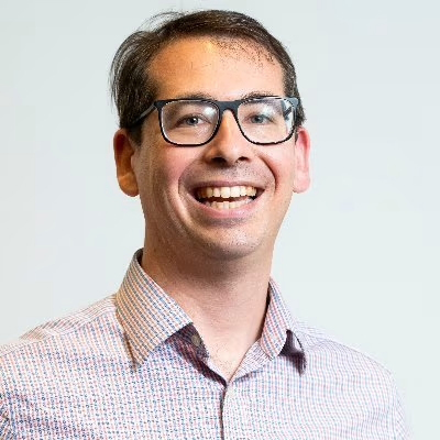 A person with glasses wearing a checkered button-down shirt, smiling broadly at the camera against a light background.