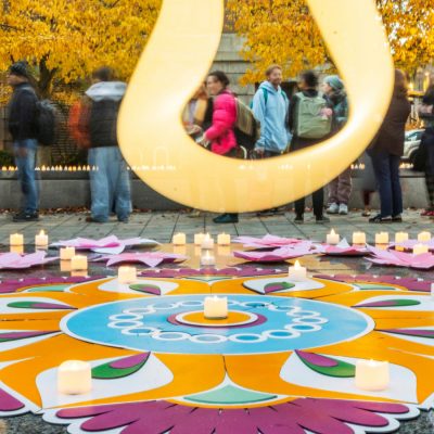 Large golden sculpture frames people in autumn plaza with colorful mandala floor decoration and lit candles surrounded by yellow fall trees.