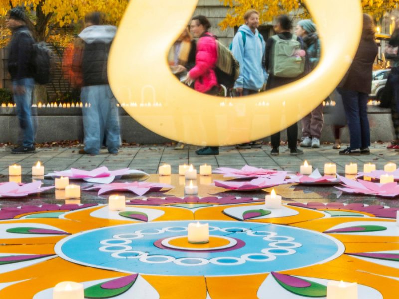 Large golden sculpture frames people in autumn plaza with colorful mandala floor decoration and lit candles surrounded by yellow fall trees.