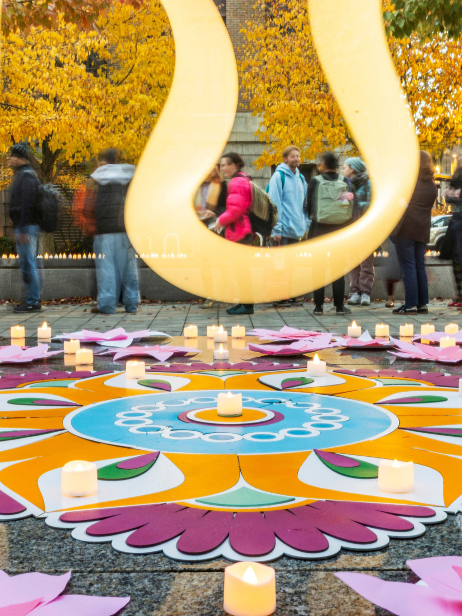 Large golden sculpture frames people in autumn plaza with colorful mandala floor decoration and lit candles surrounded by yellow fall trees.