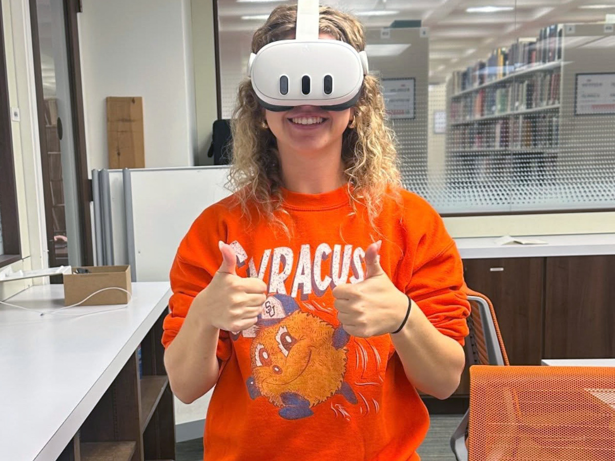 Student wearing a Syracuse sweatshirt and VR headset giving thumbs up in a classroom setting