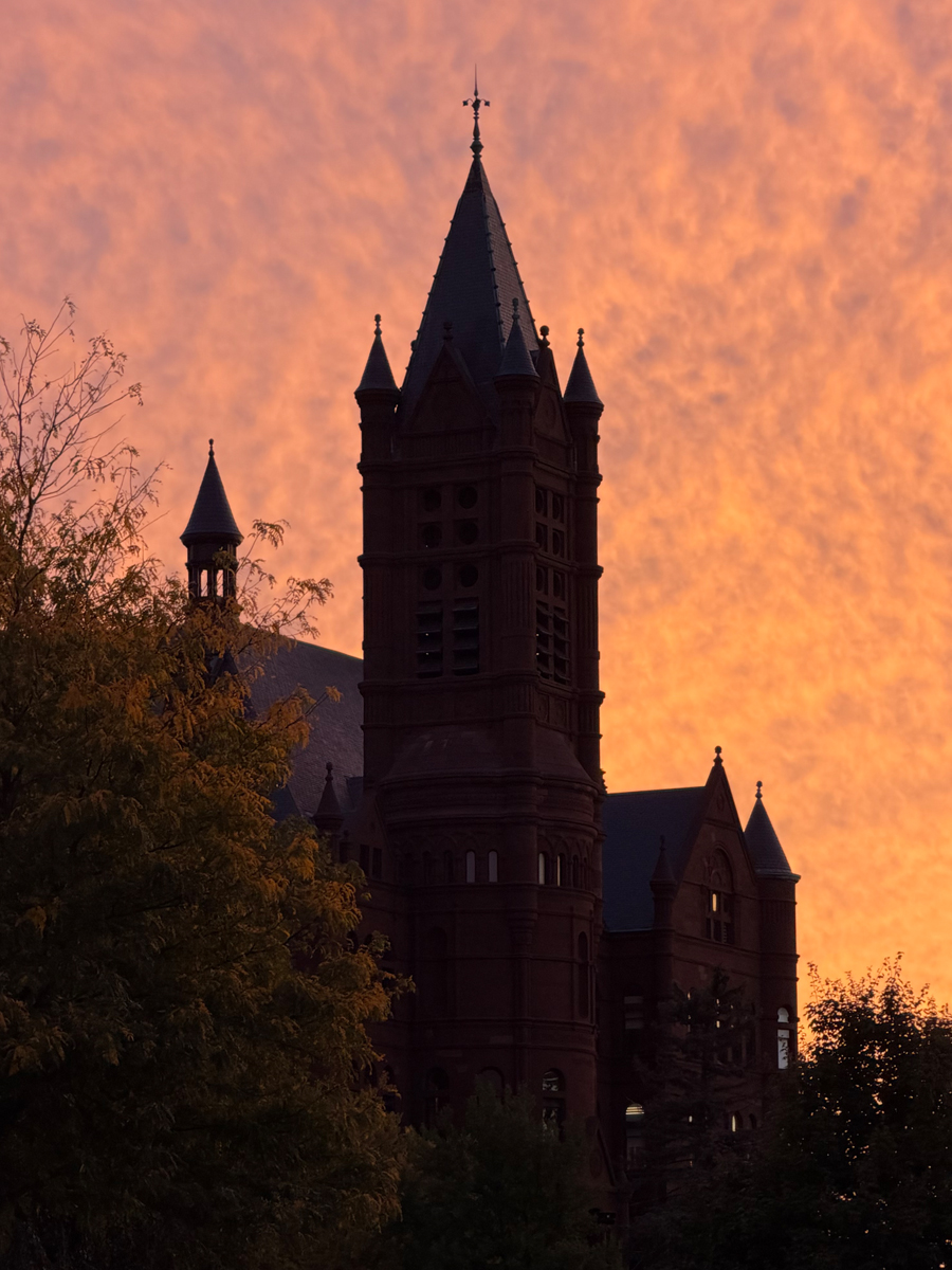 Historic Crouse College silhouetted against vibrant orange and pink sunset sky with autumn trees in foreground.