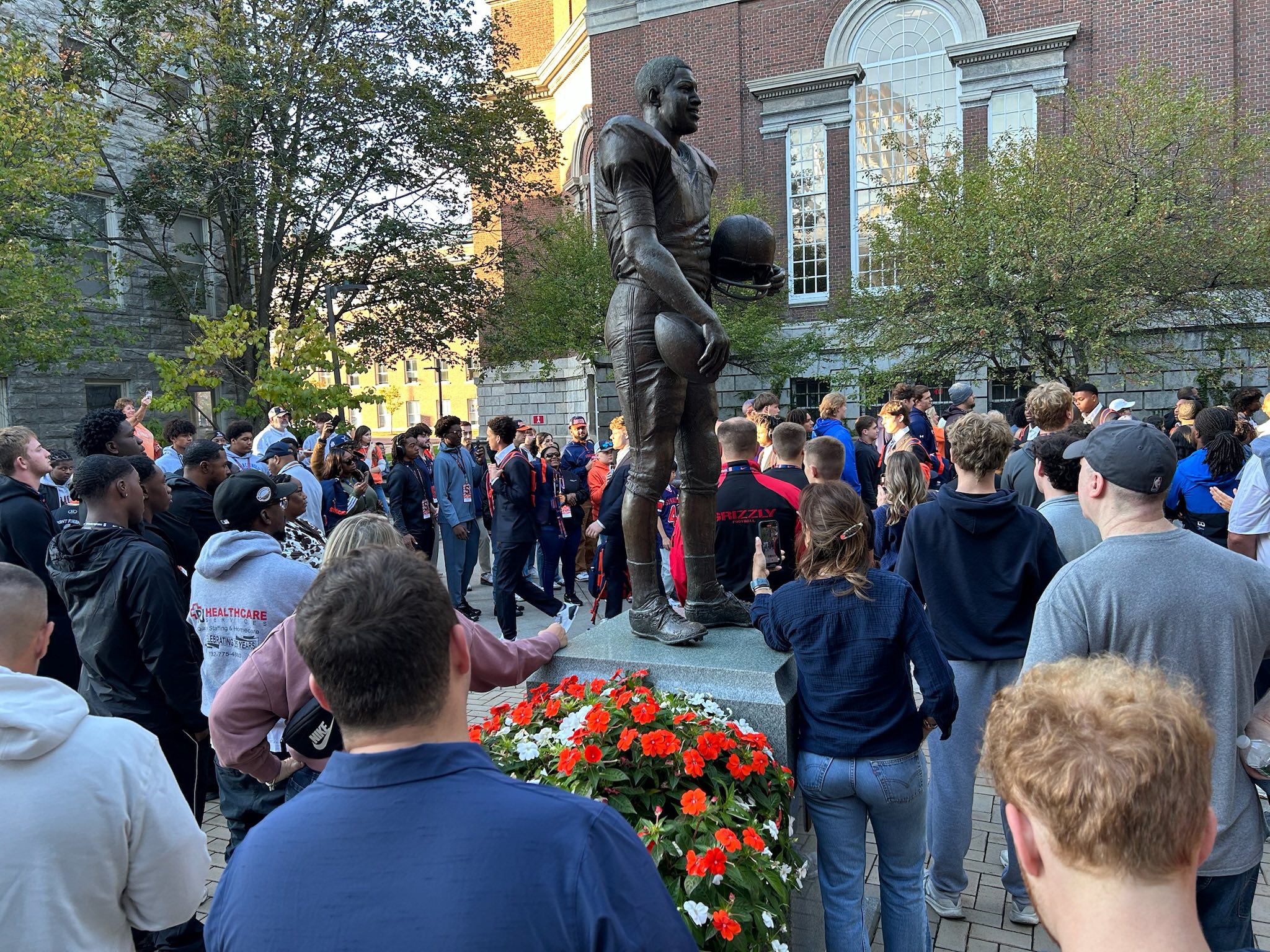 Large crowd gathered around Ernie Davis statue on campus with orange flowers at base during commemoration event