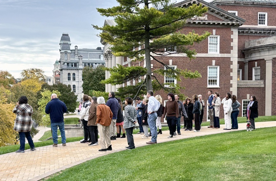 Group of people on a guided fall foliage tour near a historic red-brick building with white columns, surrounded by autumn trees.