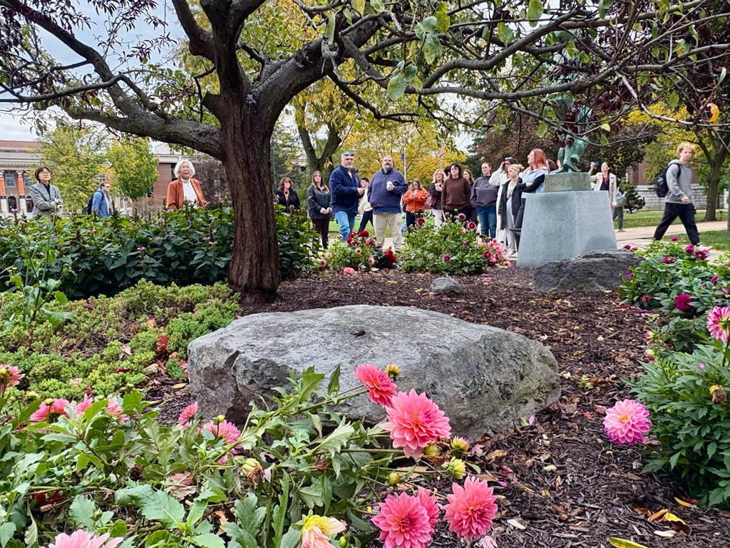 Group of people on a walking tour gathered near a large rock and pink flowers in a shaded area with statue and lush greenery in the background
